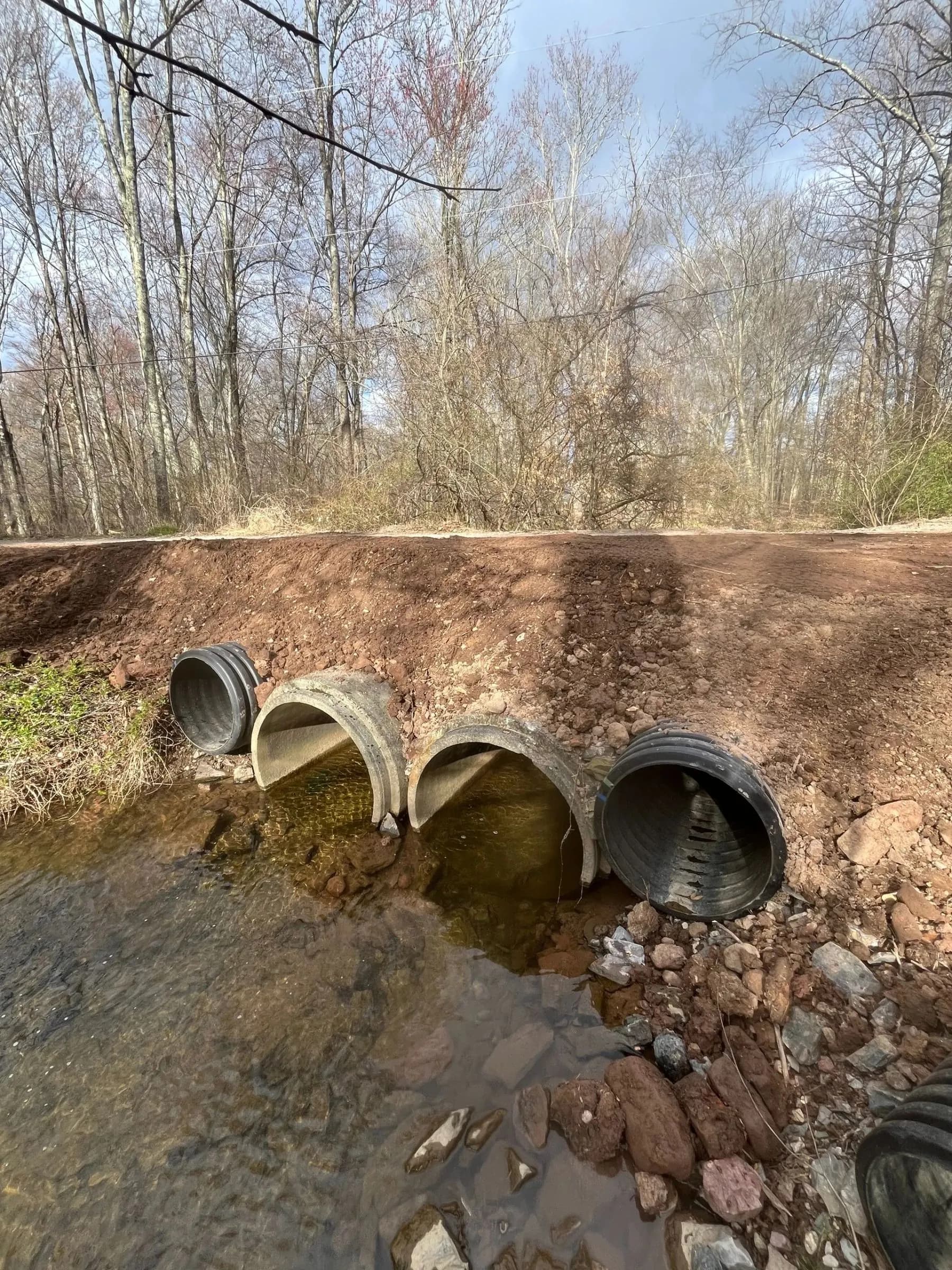 Four large drainage pipes set into a dirt bank above a rocky stream in a wooded area.