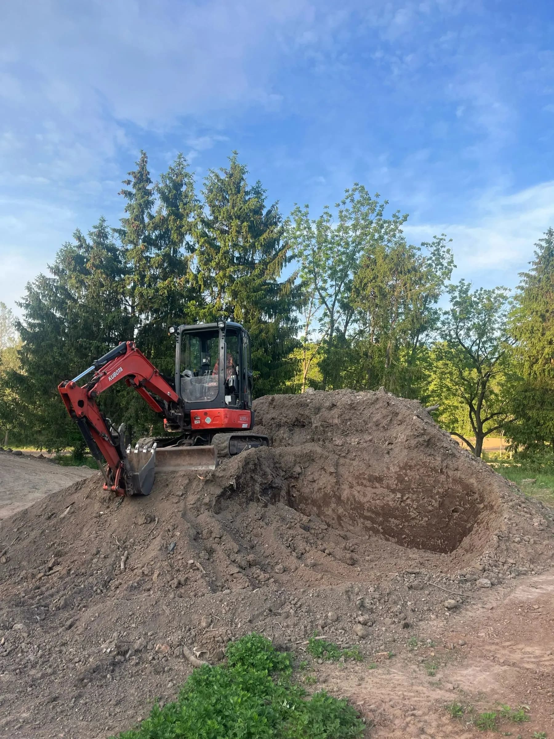 A Kubota excavator digging into a mound of soil on an open site.