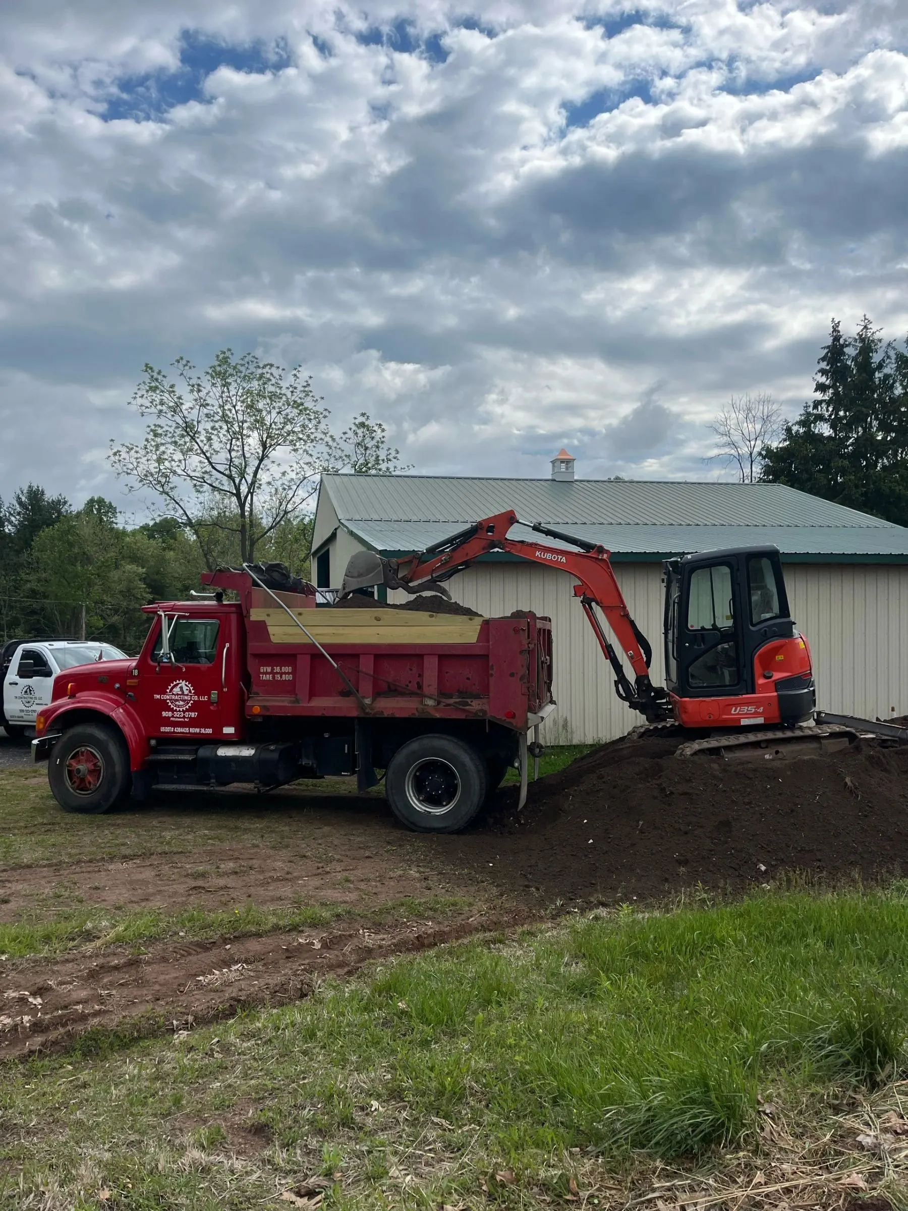 Kubota excavator loading the TM Contracting dump truck beside a farm building.