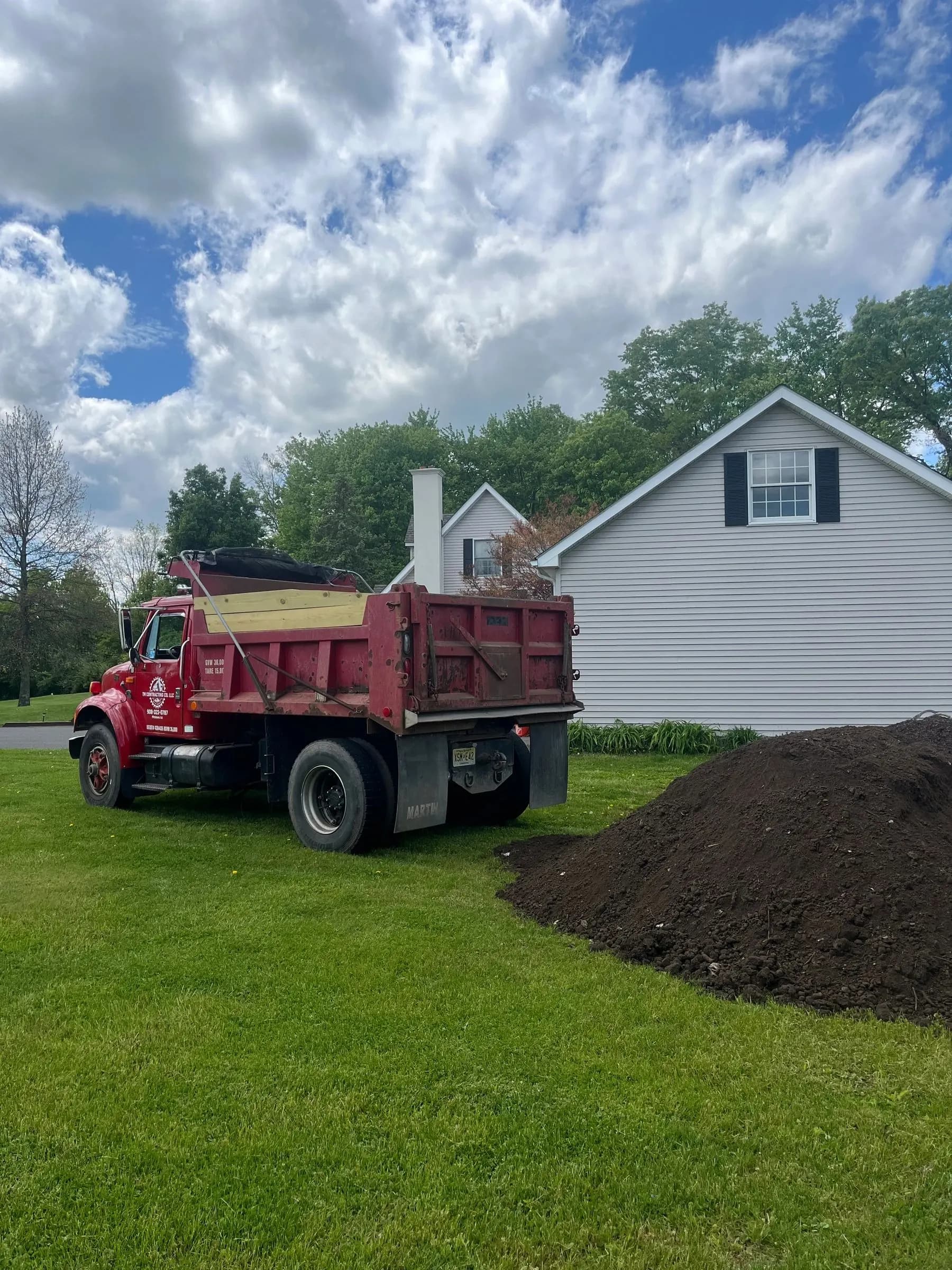 A TM Contracting dump truck that has dropped a pile of dark soil beside a house.
