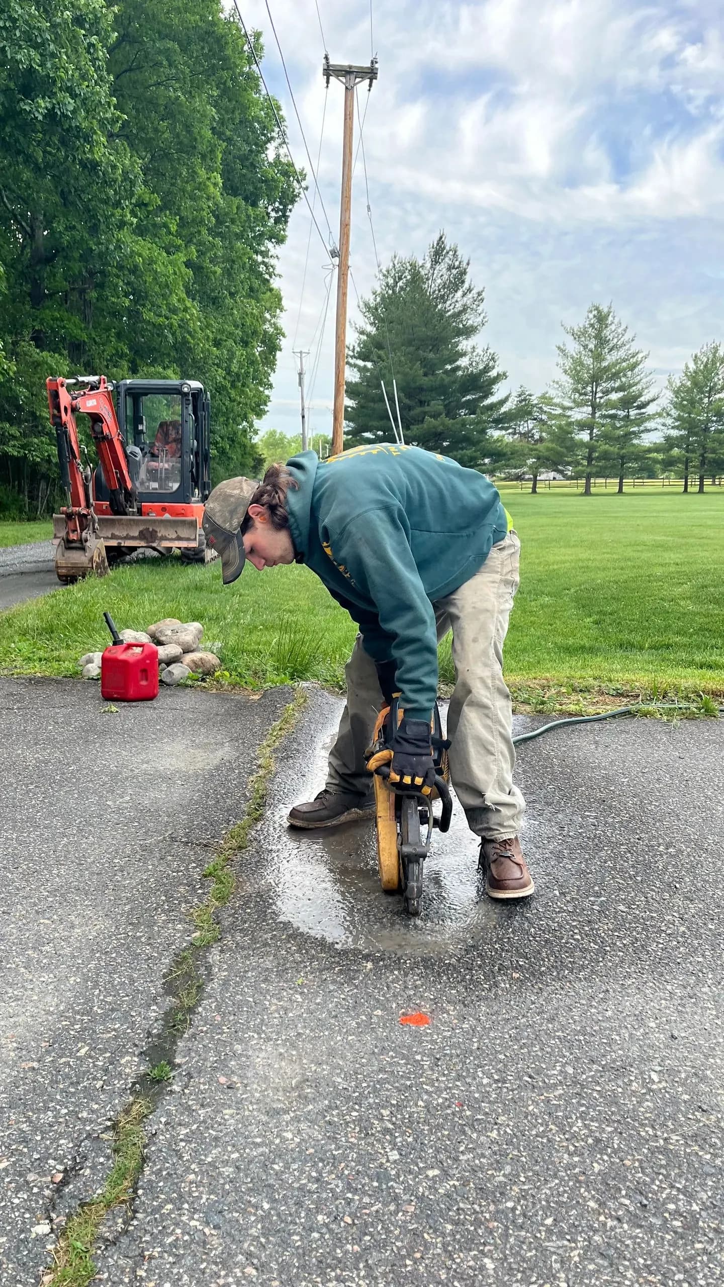 A crew member cutting asphalt with a saw, an excavator and open pasture behind him.