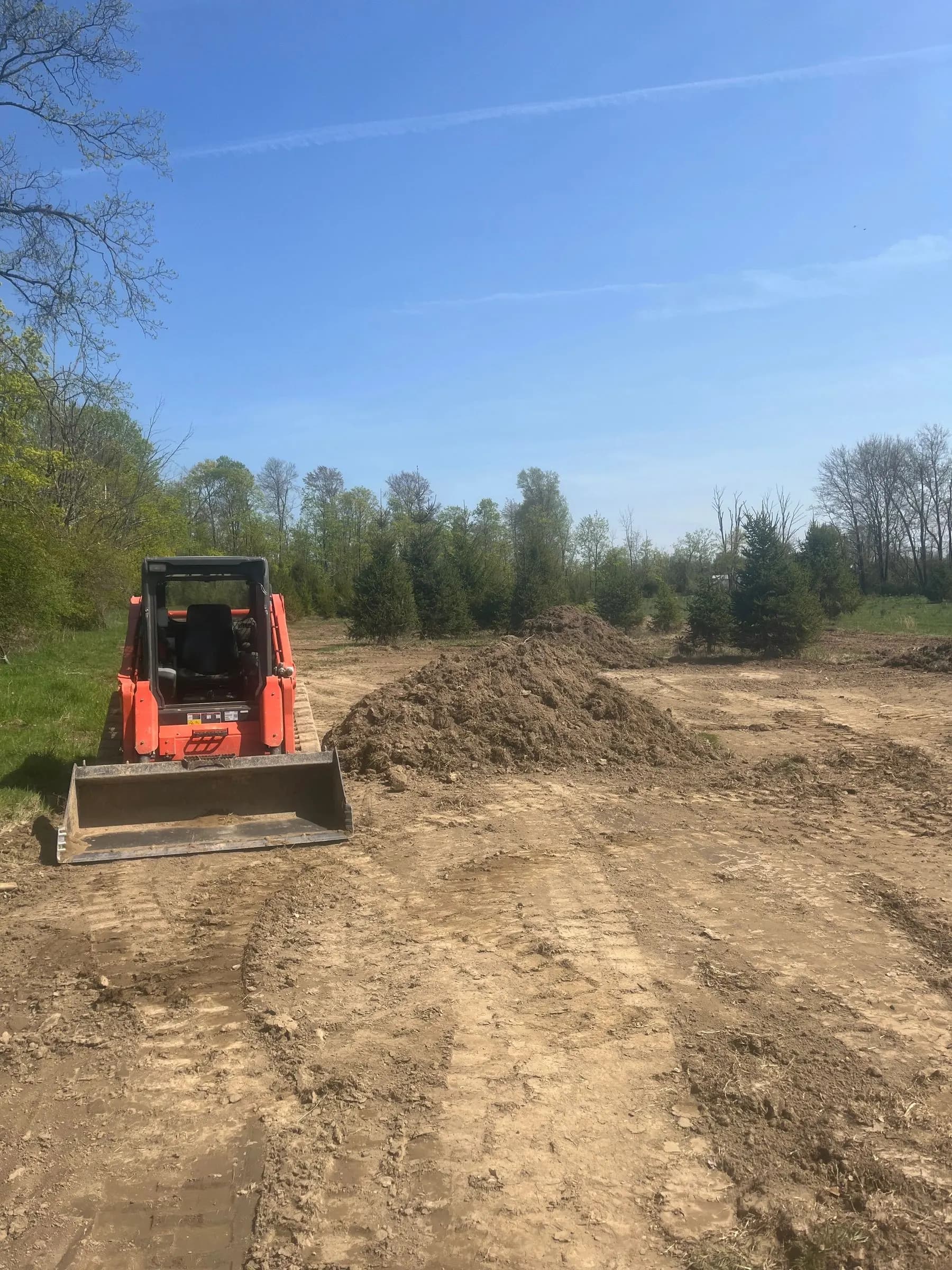 A track loader on a partially cleared field with soil piles and tree lines beyond.